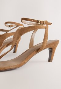 Des sandales à talons en daim marron présentent une bride fine à la cheville avec une boucle dorée, un design à bout ouvert et un talon légèrement effilé.