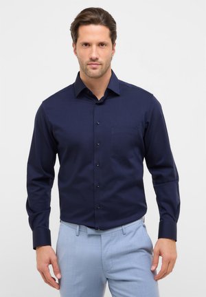 Chemise bleu marine à boutons avec manches longues, une poche poitrine simple et une texture de tissu lisse. Caractérisée par un col classique et des boutons foncés.