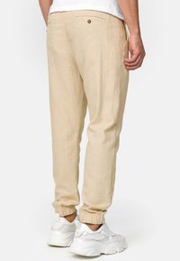 Pantalones jogger de lino beige con puños elásticos, detalles de bolsillo trasero con botón y un ajuste relajado, combinados con zapatillas deportivas blancas.