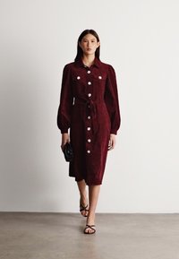 Burgundiszínű, velúr midi ruha hosszú puffujjakkal, elülső gombokkal és megkötött derék részlettel. Szandálokkal és fekete clutch-táskával párosítva.