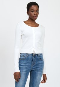 Cárdigan blanco de mangas largas con cuello redondo, cierre de botones y puños acanalados, combinado con unos jeans de mezclilla azul de cintura alta.