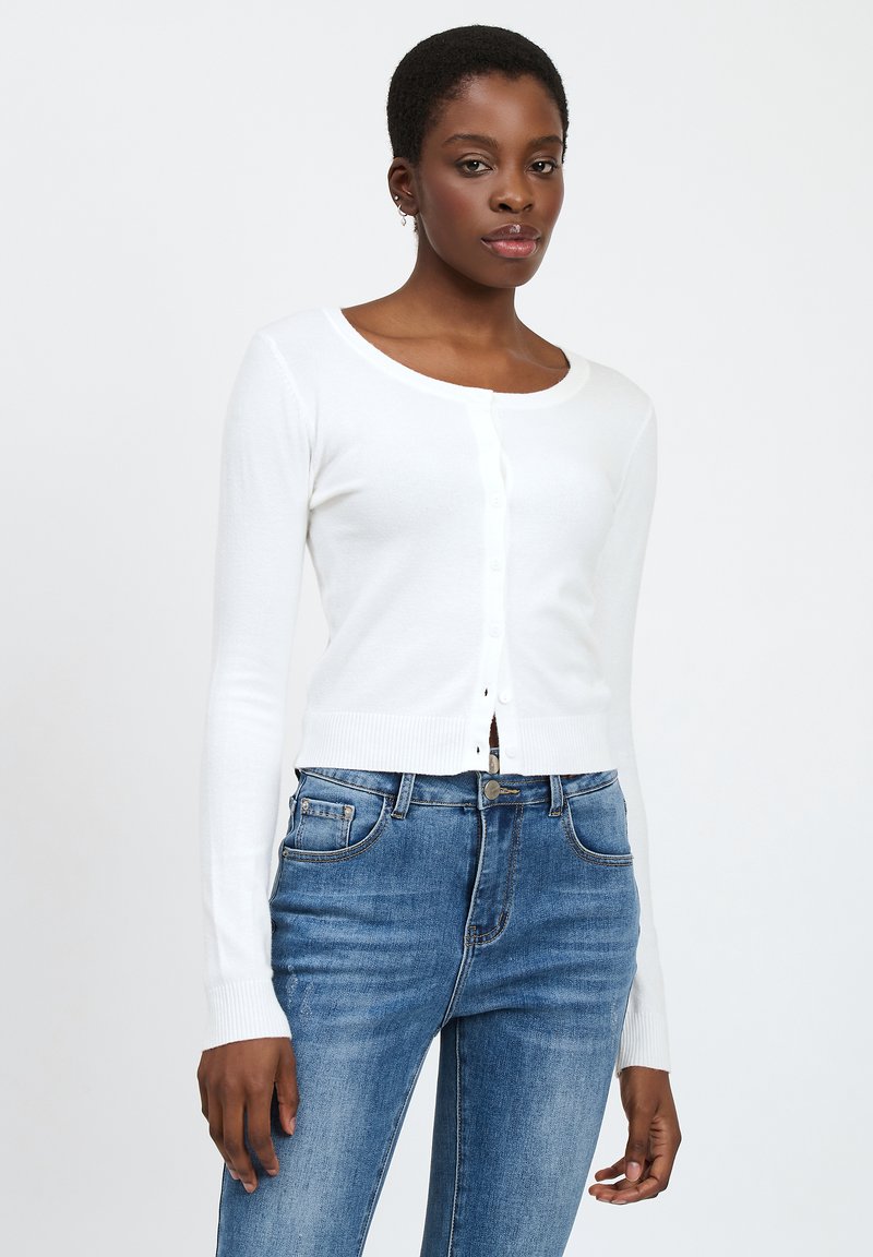 Cárdigan blanco de mangas largas con cuello redondo, cierre de botones y puños acanalados, combinado con unos jeans de mezclilla azul de cintura alta.