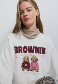 Sudadera blanca con un gráfico en color burdeos y rosa que muestra a dos perros con suéteres. La modelo también lleva un cuello alto beige debajo.