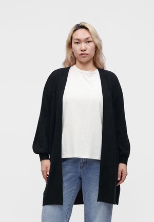 Jeune personne aux cheveux blonds porte un long cardigan noir, un t-shirt blanc et un jean bleu, debout devant un fond clair uni.