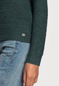 Gebreide trui in donkergroen met een gestructureerd patroon, ribgebreide manchetten en een klein zilveren label aan de zoom; gecombineerd met lichtblauwe jeans.