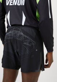 Schwarze Sportshorts mit einem geometrischen Grafikdesign in Grau, mit einem gespaltenen Saum und elastischem Bund. Das Herstellerlogo ist sichtbar.
