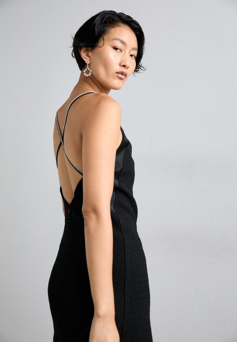 Schwarzes Kleid aus strukturiertem Stoff, mit offenem Rücken und überkreuzten Trägern. Verfügt über glänzende Akzente entlang der Ränder der Träger und eine figurbetonte Silhouette.