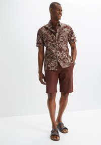 Camicia a maniche corte marrone con un leggero motivo a foglie di palma, abbinata a pantaloni corti marroni coordinati e sandali grigi con fibbie. Tessuto liscio.