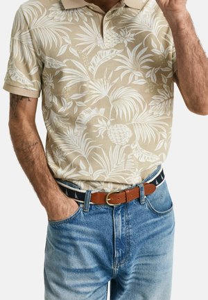 Polo beige avec un imprimé blanc de feuilles tropicales et d'ananas, associé à un jean bleu délavé et une ceinture en cuir marron.