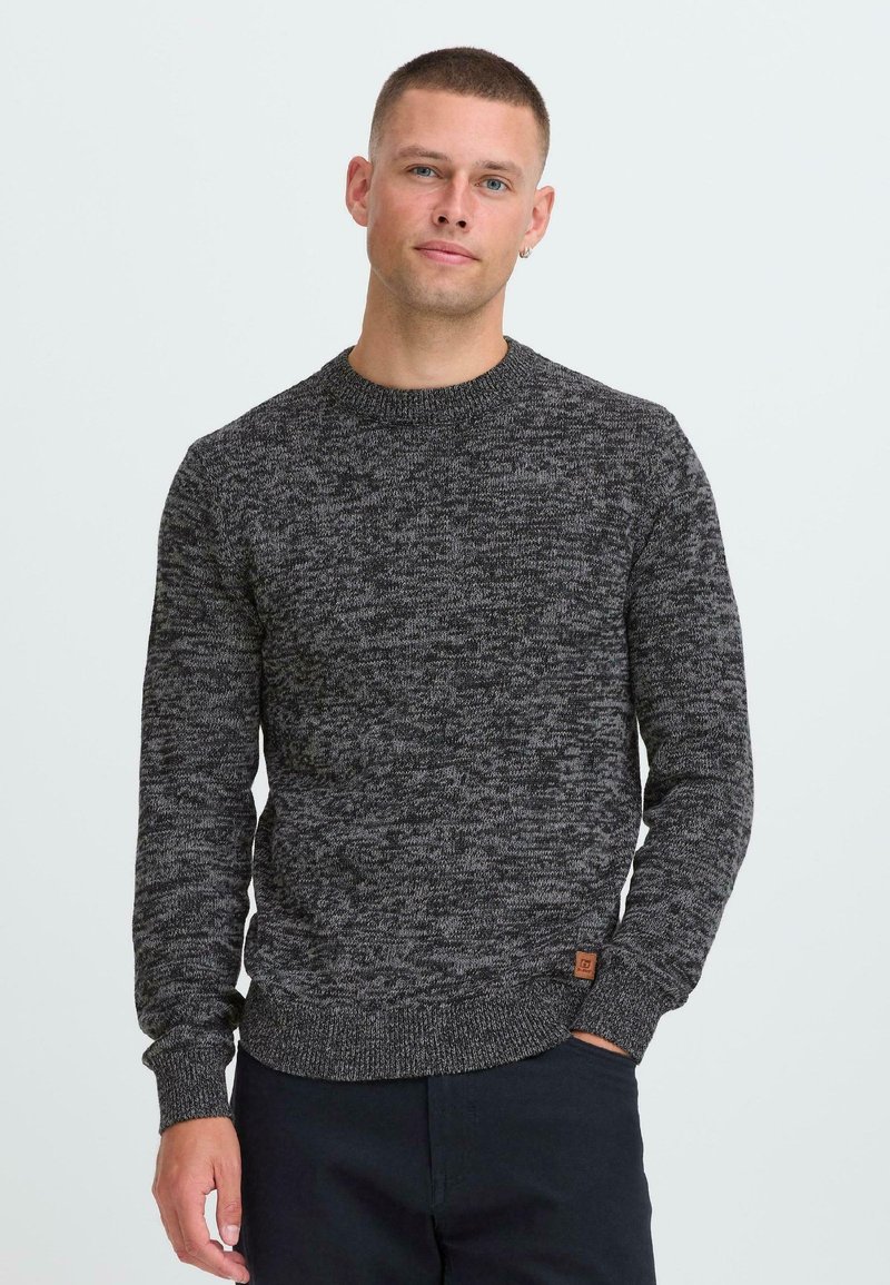 Man met kort haar in een donkergrijze gemarmerde gebreide trui en zwarte broek, staand met één hand in de zak tegen een lichte achtergrond.