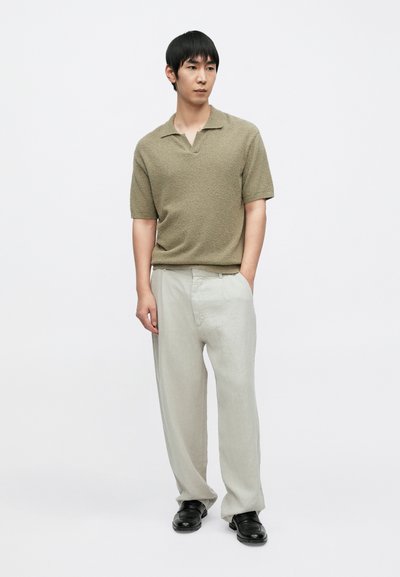 Πόλο πουκάμισο κοντομάνικο με υφή σε ελιά πράσινη απόχρωση, συνδυασμένο με ανοιχτό γκρι παντελόνι σε άνετη γραμμή και μαύρα παπούτσια, στέκεται μπροστά σε απλό φόντο.