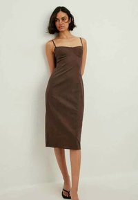 Brun midi-kjole med en tettsittende silhuett, tynne stropper og en jevn tekstur. Har rett utringning og subtil detaljer på overdelen.
