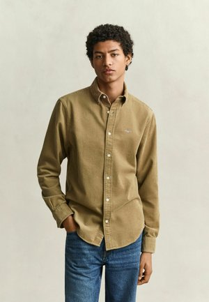 Jeune homme aux cheveux bouclés portant une chemise beige boutonnée et un jean bleu, debout avec une main dans la poche devant un fond uni.