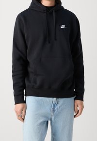 Svart huvtröja med framficka, dragsko på huvan och vit Nike-logotyp på bröstet. Mjuk tyg och avslappnad passform. Ljusblå jeans.