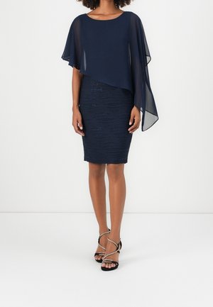 Frau trägt ein marineblaues One-Shoulder-Kleid mit transparentem Überzug und schwarzen Riemchen-High-Heel-Sandalen.