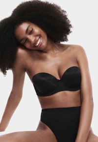 Femme souriante aux cheveux bouclés portant un soutien-gorge noir sans bretelles et une culotte noire taille haute, sur un fond blanc uni.