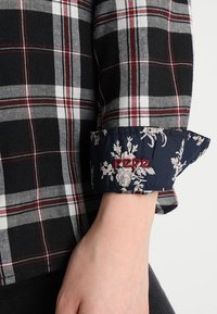 Black, white, and red plaid shirt sleeve rolled up to show navy cuff with white floral pattern and red embroidered "pepe" text.