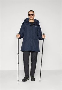 Γυναίκα με ναυτικό μπουφάν, μαύρα παντελόνια, μποτάκια πεζοπορίας και γυαλιά ηλίου, κρατώντας μαύρα πεζοπορικά μαστίγια, στέκεται μπροστά σε απλό φόντο.