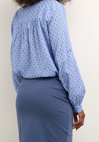 Chemise bleu clair à petits pois bleu foncé, avec manches longues et dos froncé ; associée à une jupe bleue ajustée.