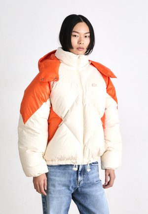 Chaqueta acolchada en crema y naranja, con un diseño geométrico acolchado, cierre de cremallera y dobladillo ajustable con cordón. Logo icónico en el pecho.
