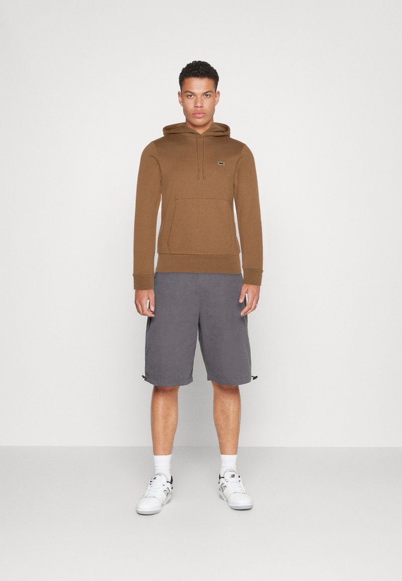 Bruine hoodie met een voorzak en trekkoord, gecombineerd met grijze shorts en witte sneakers. Eenvoudig ontwerp met een gestructureerde afwerking.