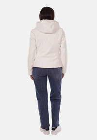 Giacca bianca con cappuccio, dal texture morbido, design aderente e vita leggermente ristretta. Indossata con jeans blu e sneakers bianche.