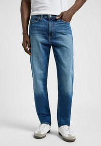 Jeans de mezclilla azul con un diseño de pierna recta, que presentan desvanecimiento cerca del muslo, estilo clásico de cinco bolsillos y cierre de botón de metal.