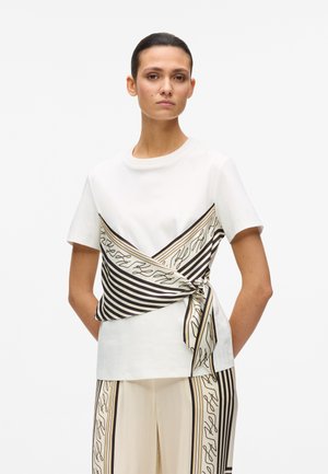 Kvinde iført hvid kortærmet top med sort og beige stribet mønstret tørklæde bundet om taljen, matchende mønstrede bukser med vide ben.