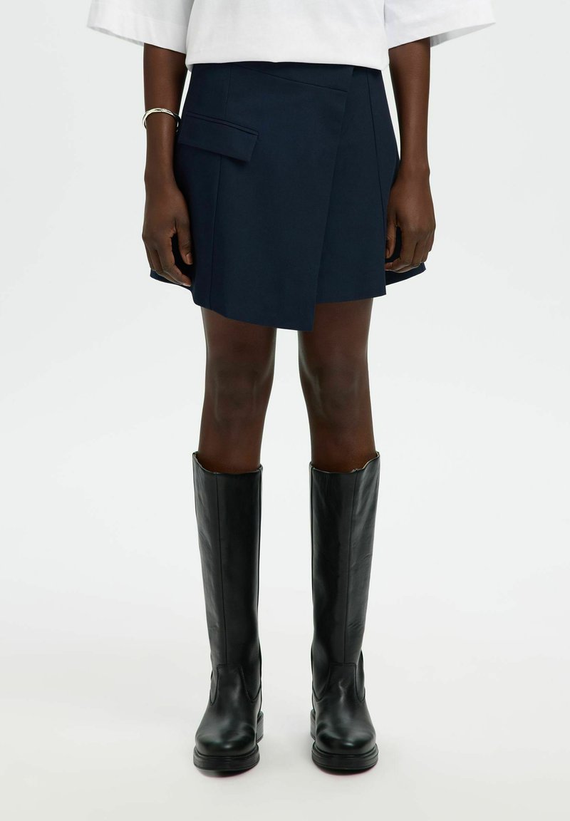 Asmuo, dėvintis tamsiai mėlyną mini sijoną su asimetriška kišene ir aukštais juodos odos batais, stovintis priešais šviesią foną.