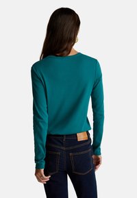 Türkises Langarmshirt mit rundem Halsausschnitt, kombiniert mit dunklen Jeans aus Denim mit Kontrastnähten und Gesäßtaschen. Keine sichtbaren Logos.