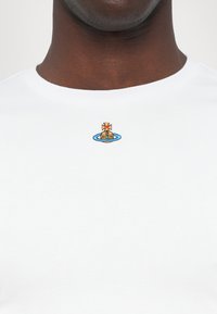 T-shirt en coton blanc avec un col rond, présentant un petit motif brodé d'un bateau et d'une croix dans les couleurs bleu, or et rouge.