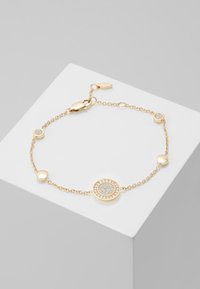 Pulsera de cadena en tono dorado que presenta encantos circulares, uno de ellos con una superficie texturizada rodeado de pequeñas piedras y acentos redondeados lisos.