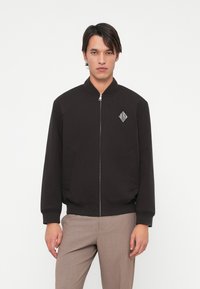 Mustmustaga must jacket, mille avaga, soonikest ja kahe külje taskuga. Vasakul rinnal on väike valge geomeetriline logo.