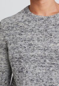 Pull en tricot gris avec un col rond. La texture présente un motif moucheté de gris plus foncé et de noir.