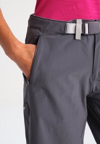 Grå lätta byxor med en slät textur, ett bälte med en silverfärgad spänne och sidofickor med dragkedjor.