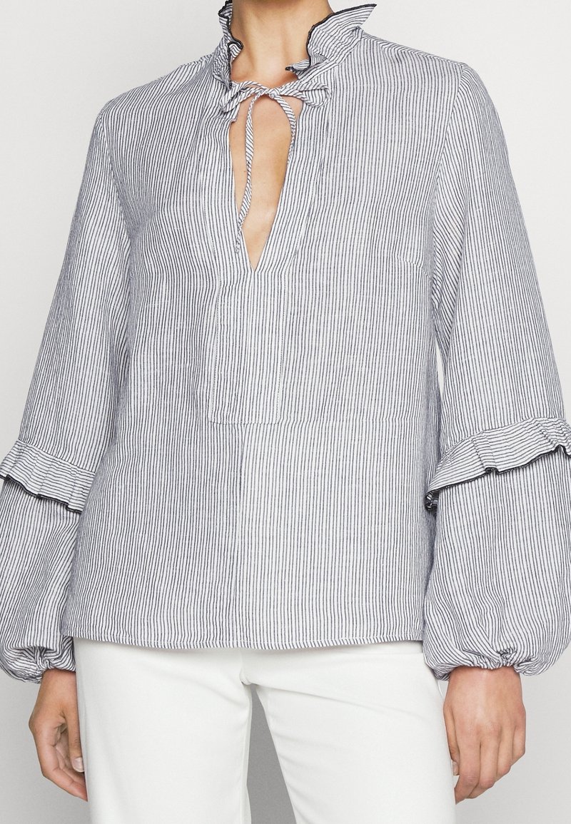 Blouse blanche à manches longues avec fines rayures verticales noires, col volanté noué d'un nœud et manches bouffantes avec détail à volants.