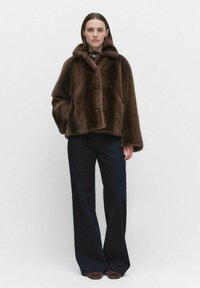 Brązowa kurtka z sztucznego futra z kołnierzem, trzema guzikami i luźnym krojem. W połączeniu z ciemnymi spodniami o szerokich nogawkach i brązowymi butami typu slip-on.