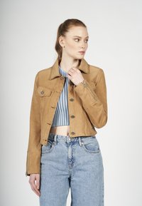 Lichtbruine suède jack met een kraag, voorzakken en ritsdetails. Gecombineerd met een blauwe geribbelde crop top en lichte denim jeans.