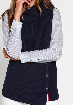 Marineblauer gestrickter ärmelloser Pullover mit einem Kragen aus Löchern, der seitliche Knöpfe und einen geschichteten Saum über einem gestreiften Hemd aufweist.