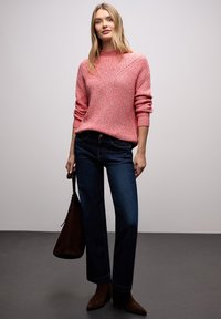 Femme portant un pull en tricot rose, un jean bleu foncé, des bottes marron, et tenant un sac à main marron foncé sur un fond uni.