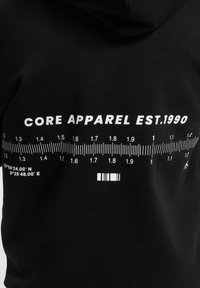 Svart hettegenser med hvit tekst "CORE APPAREL EST.1990" og et linjal-illustrasjon med koordinater og strekkode, laget av myk stoff.