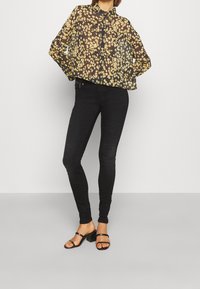 Blusa nera trasparente con motivo floreale giallo, colletto con bottoni e maniche lunghe, abbinata a jeans skinny neri e tacchi neri con cinturini.