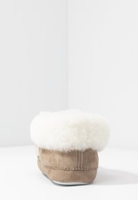 Brown suede slipper with white fluffy lining, featuring a rounded shape and stitched accent. Subtle label detail on the side.