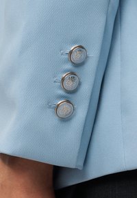 Manche de blazer bleu clair avec trois boutons émaillés bleus à bordure argentée ornés de dessins complexes de blasons.