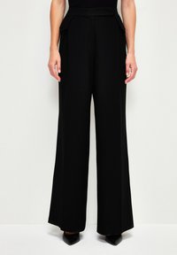 Zwarte wijde broek van soepel stof met een gestructureerde tailleband en zijplooien, gecombineerd met puntige zwarte hakken.