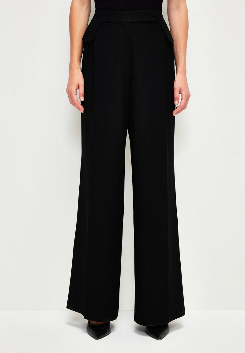 Zwarte wijde broek van soepel stof met een gestructureerde tailleband en zijplooien, gecombineerd met puntige zwarte hakken.
