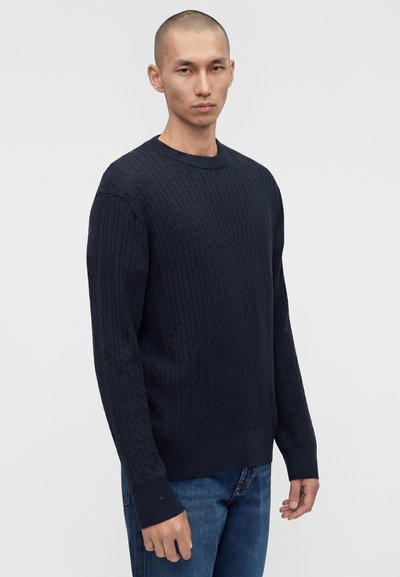 Άνδρας με σκούνο μπλε πουλόβερ με ανάγλυφο σχέδιο και μπλε τζιν, στέκεται μπροστά σε απλό ανοιχτογρίζο φόντο.