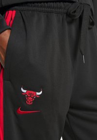 Sorte træningsbukser med røde sidelister, med et rødt og hvidt broderet tyr-logo og en lille rød Nike swoosh. Justerbar talje.