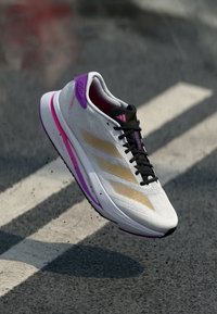 Scarpa da corsa con tomaia in rete bianca, strisce dorate, accenti viola, lacci neri e una suola ammortizzata, posizionata sopra una superficie texturizzata.