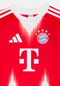 Crvena nogometna majica s bijelim okruglim izrezom, Adidas prugama, velikim bijelim logotipom sponzora i emblemom FC Bayerna s akcentima u obliku zvijezda.